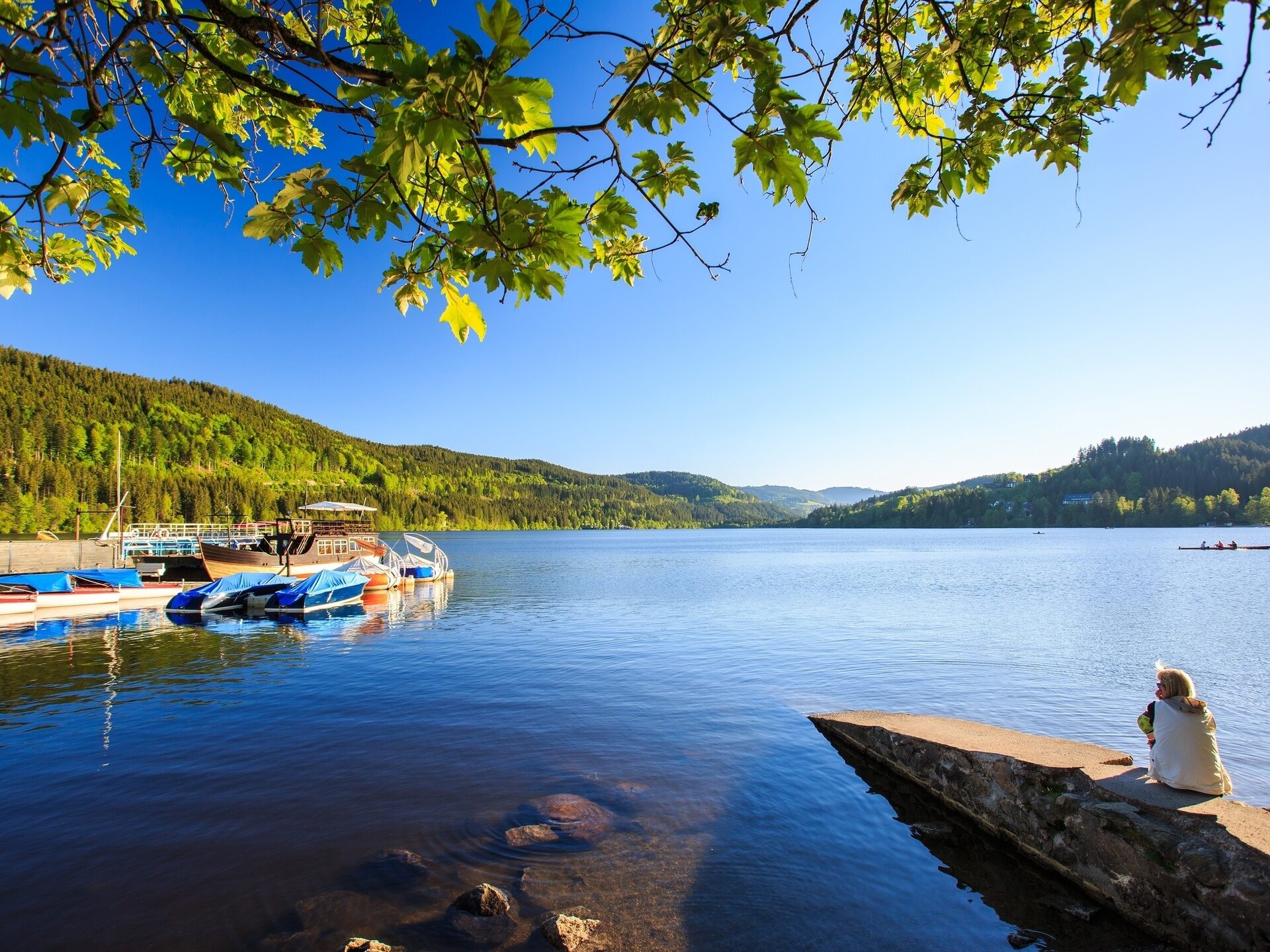 Eine Person sitzt an einem See der umrahmt von H&uuml;geln ist.