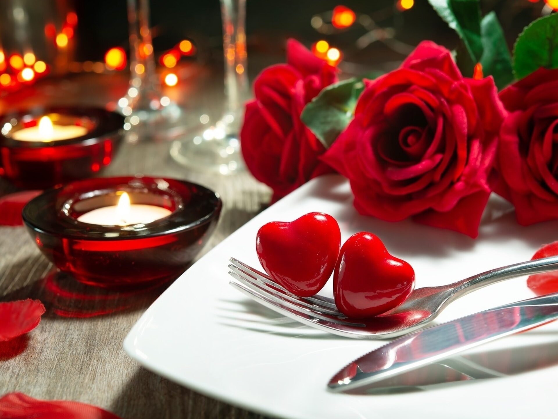 Auf einem mit roten Rosen und Kerzen dekorierter Tisch steht ein Teller mit Besteck auf dem rote Herzen liegen.